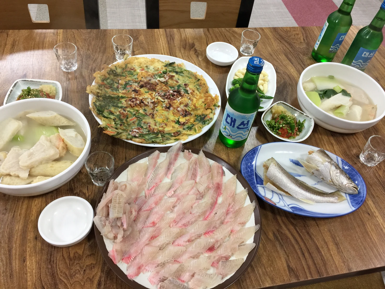 木の卓に刺身の皿、チヂミ、汁物、焼き魚が一緒に並べられている。